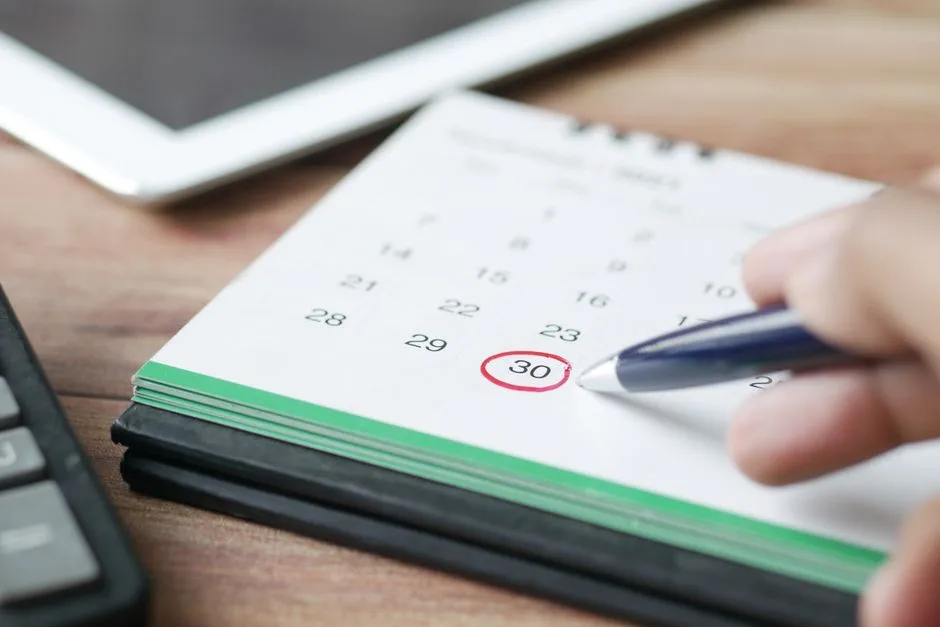 Mână marcând cu pixul o dată încercuită pe un calendar de birou, lângă un calculator de birou