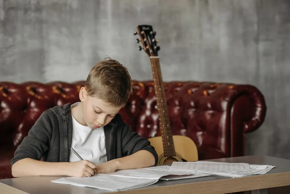 Băiat scriind la masă, cu o chitară acustică sprijinită de canapea în spate