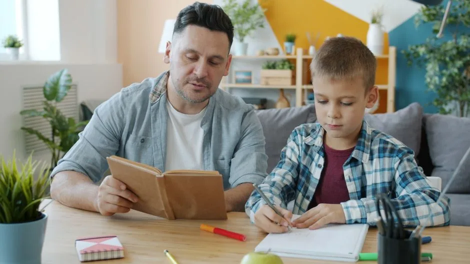 Tată citind o carte la masă lângă un băiat care scrie într-un caiet
